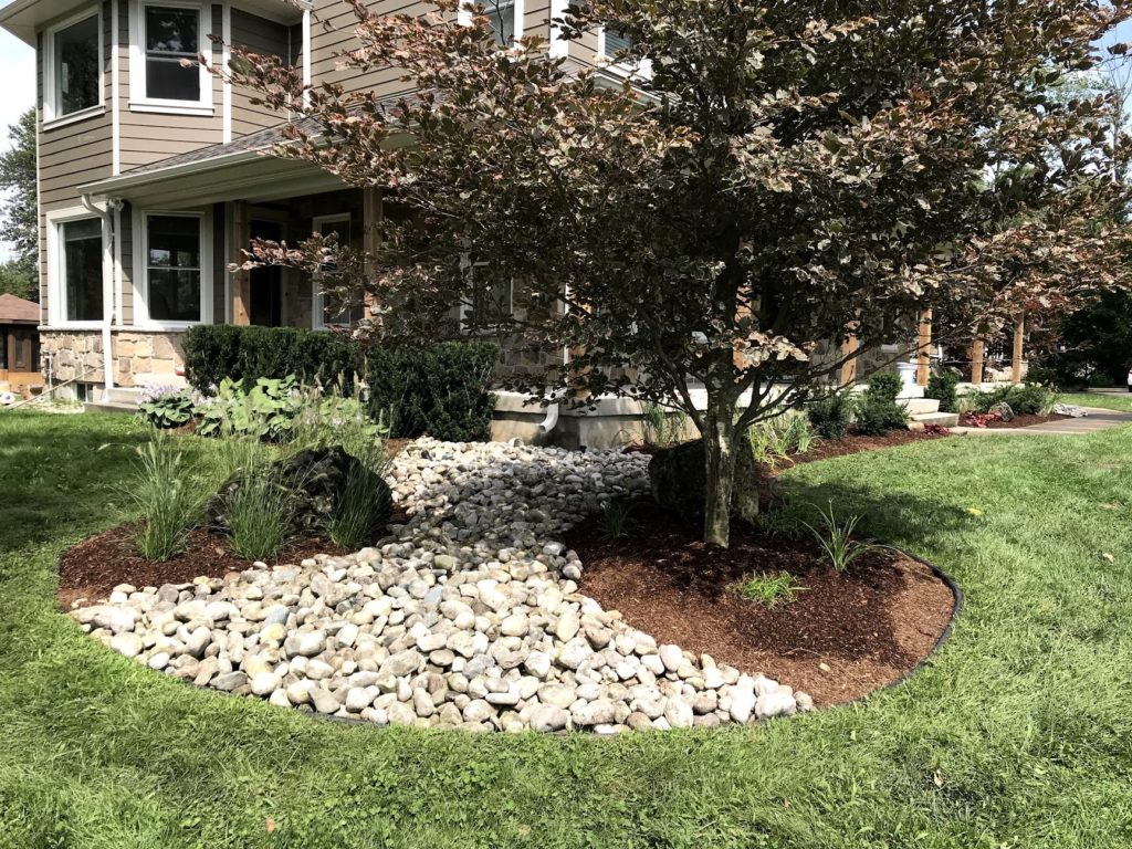 A garden in front of a house with a tree and some small rocks.