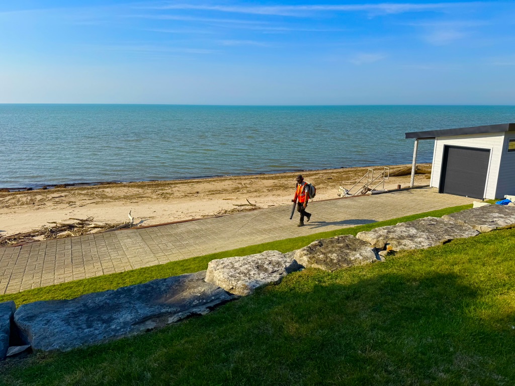 A man walking along a path beside a beach, using a leaf blower.
